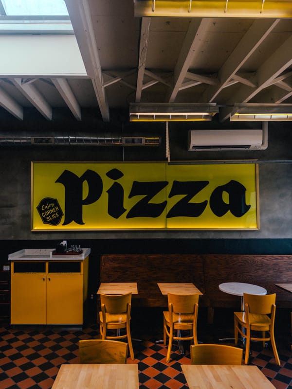 Interior of a pizza place, 5 chairs and tables are visible, a trash bin to the left, and a big yellow sign that says "PIZZA" in blackletter